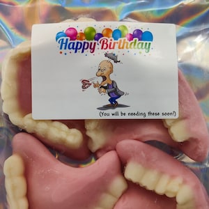 May include: A clear plastic bag containing four pink and white gummy dentures. A white card with the text "Happy Birthday" and a cartoon image of a person with dentures is attached to the bag.