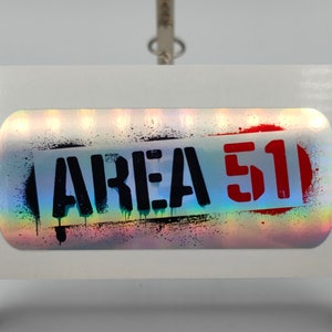 Może przedstawiać: Holograficzna naklejka z tekstem "AREA 51" w czarnych i czerwonych literach. Naklejka ma tęczowy, opalizujący tło.