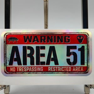 Może przedstawiać: Holograficzna naklejka z czerwonym tłem i czarnym tekstem, który brzmi "WARNING AREA 51 NO TRESPASSING RESTRICTED AREA". Naklejka zawiera grafikę UFO i głowy kosmity.