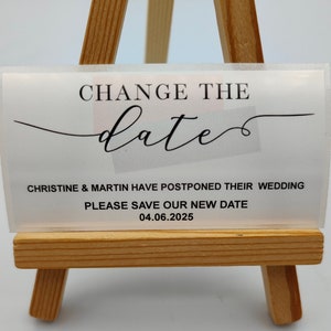 Puede incluir: Una tarjeta blanca con texto negro en un caballete de madera. La tarjeta dice "Change the date" con una fuente de letra cursiva decorativa. El texto debajo dice "Christine & Martin han pospuesto su boda. Por favor, guarde nuestra nueva fecha 04.06.2025."