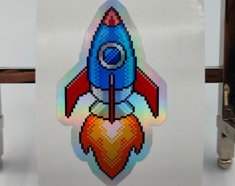 Könnte beinhalten: Ein holografischer Aufkleber mit einer pixelartigen Rakete mit roten und blauen Akzenten und einer orangefarbenen Flamme. Die Rakete befindet sich auf einem weißen Hintergrund.
