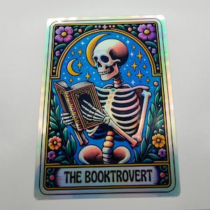 以下が含まれることがあります： 三日月と星を背景に、本を読んでいる骸骨が描かれたホログラフィックステッカー。ステッカーの下部に「THE BOOKTROVERT」というテキストがあります。