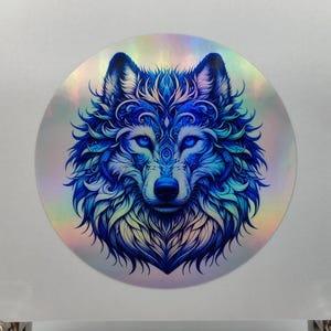以下が含まれることがあります： 複雑なディテールが施された青いオオカミが描かれた、虹色の丸いステッカー。オオカミの目は明るい青で、毛皮は渦巻く模様で飾られています。
