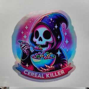 May include: A holographic sticker featuring a skeleton wearing a black hooded robe and holding a bowl of cereal. The text "Cereal Killer" is written in pink on a banner below the skeleton.