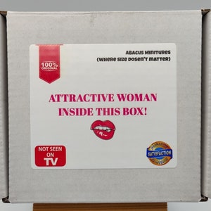 May include: A white cardboard box with the text "Attractive Woman Inside This Box!" and a red lip print. The box also has a blue and gold sticker that says "Guaranteed Satisfaction".