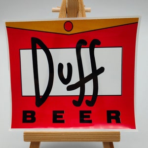 May include: A red and gold sticker with the words "Duff Beer" in black lettering. The sticker is on a small wooden easel.