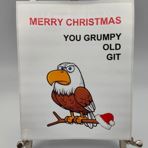 Può includere: Un biglietto di Natale umoristico con un'aquila calva dei cartoni animati con un cappello di Babbo Natale su un ramo. Il biglietto presenta il testo "MERRY CHRISTMAS YOU GRUMPY OLD GIT" in rosso e nero su sfondo bianco.