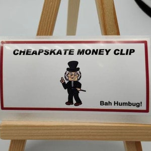 Puede incluir: Una pegatina blanca con borde rojo en un caballete de madera. La pegatina presenta una ilustración de dibujos animados de un hombre con un sombrero de copa y un bastón. El texto en la pegatina dice "Cheapskate Money Clip" y "Bah Humbug!"