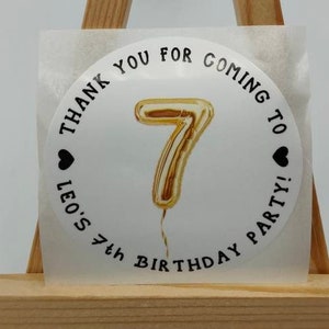 Pode incluir: Etiqueta redonda branca com texto preto que diz "Obrigado por vir à festa de aniversário de 7 anos do Leo!" com um balão número 7 dourado no centro.
