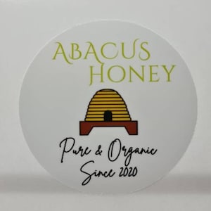 May include: White round sticker with the text "Abacus Honey" in green lettering. A cartoon beehive with a brown base is in the center of the sticker. The text "Pure & Organic Since 2020" is below the beehive.