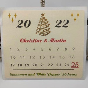 Op de afbeelding: Een witte kaart met een gouden kerstboomontwerp en de tekst "20 Christine & Martin 22" en een kalender met de nummers 1 tot en met 25. De tekst "Kaneel en witte peper | 30 uur" staat onderaan de kaart.