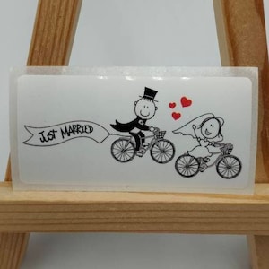 May include: A white sticker with a cartoon illustration of a bride and groom riding bicycles. The groom is wearing a top hat and the bride has a veil. A banner above them reads "Just Married".