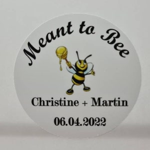 May include: A white round sticker with a cartoon bee holding a honey dipper. The text on the sticker reads "Meant to Bee" with the names "Christine + Martin" and the date "06.04.2022" below the bee.