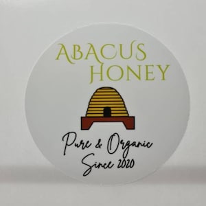 May include: A white round sticker with the text "Abacus Honey" in green letters. A cartoon image of a beehive with a brown base and yellow honeycomb is in the center. The text "Pure & Organic Since 2020" is below the beehive.