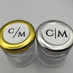 Puede incluir: Dos pequeños frascos de vidrio con tapas. Un frasco tiene una tapa dorada con una etiqueta blanca que dice "C/M". El otro frasco tiene una tapa plateada con una etiqueta blanca que dice "C|M".