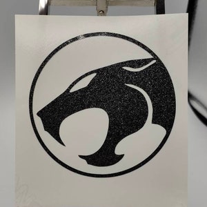 Könnte beinhalten: Schwarz-weißer Vinyl-Aufkleber mit dem ThunderCats-Logo. Das Logo zeigt einen stilisierten schwarzen Pantherkopf in einem Kreis.