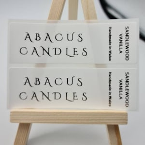 Könnte beinhalten: Zwei transparente Etiketten mit schwarzem Text auf einem Holzständer. Die Etiketten lauten "Abacus Candles" und "Sandalwood Vanilla Handmade in Wales".
