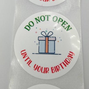 Peut inclure: Autocollant rond blanc avec du texte vert et rouge qui dit "Ne pas ouvrir avant votre anniversaire". Une illustration de dessin animé d'une boîte cadeau avec un ruban rouge est au centre de l'autocollant.