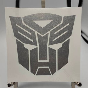 Könnte beinhalten: Ein silberner Glitzer-Vinyl-Aufkleber mit dem Autobot-Logo der Transformers. Das Logo ist ein stilisiertes Robotergesicht mit einer sechseckigen Form.
