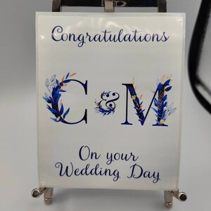 May include: A white card with blue and gold floral designs and the letters "C & M" in a blue and gold font. The card reads "Congratulations" and "On your Wedding Day".