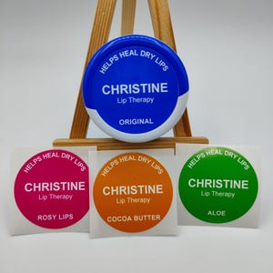 Puede incluir: Un envase de bálsamo labial azul y blanco con el texto "CHRISTINE Lip Therapy" y las palabras "HELPS HEAL DRY LIPS". Debajo hay tres pegatinas redondas en rosa, naranja y verde, cada una con el mismo texto y nombres de sabores.