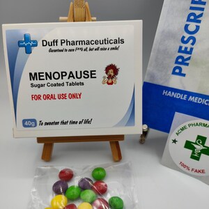 May include: A white box with a blue cross and the text "Duff Pharmaceuticals" and "Menopause Sugar Coated Tablets" with a cartoon woman on the box. The box is sitting on a wooden easel. A bag with the text "PRESCRIPT" and "HANDLE MEDIC" is in the background. A clear plastic bag with colourful candy coated chocolate pieces is in the foreground. A sticker with a green cross and the text "ACME PHARMA" and "100% FAKE" is on a white surface.