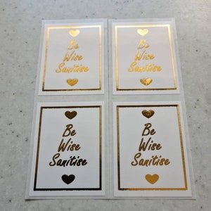 Op de afbeelding: Vier witte vierkante stickers met gouden randen en harten. Elke sticker heeft de tekst "Be Wise Sanitise" in gouden letters.
