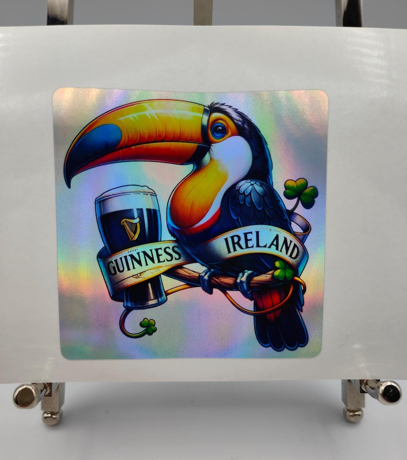 Op de afbeelding: Een toekan met een kleurrijke snavel houdt een glas Guinness-bier vast. De toekan zit op een tak met een banner waarop "Ierland" en "Guinness" staat.