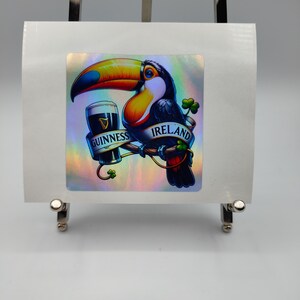 Op de afbeelding: Een kleurrijke toekan die een glas Guinness-bier vasthoudt met de tekst "IRELAND" en "GUINNESS" op een holografische sticker.