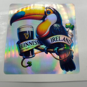 Op de afbeelding: Een toekan met een blauwe en gele snavel houdt een glas Guinness-bier vast. De toekan zit op een tak met een banner waarop "GUINNESS IERLAND" staat.