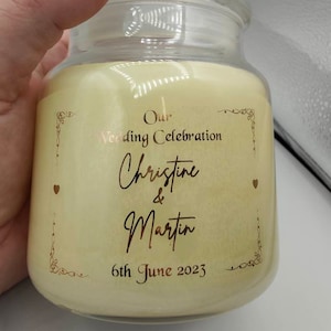 Puede incluir: Una vela en tarro de cristal transparente con cera color crema. La etiqueta presenta texto dorado que dice "Our Wedding Celebration Christine & Martin 6th June 2023". También hay detalles y corazones dorados decorativos.