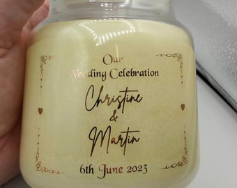 May include: A clear glass jar candle with a cream-colored wax. The label features gold text that reads "Our Wedding Celebration Christine & Martin 6th June 2023". Decorative gold accents and hearts are also present.
