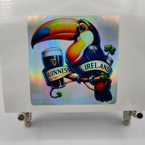 Op de afbeelding: Een holografische sticker met een toekan die een glas Guinness-bier vasthoudt. De toekan zit op een tak met een klavertjevier en het woord "IRLAND" op de tak. De sticker bevat ook de woorden "GUINNESS" en een klavertjevier.