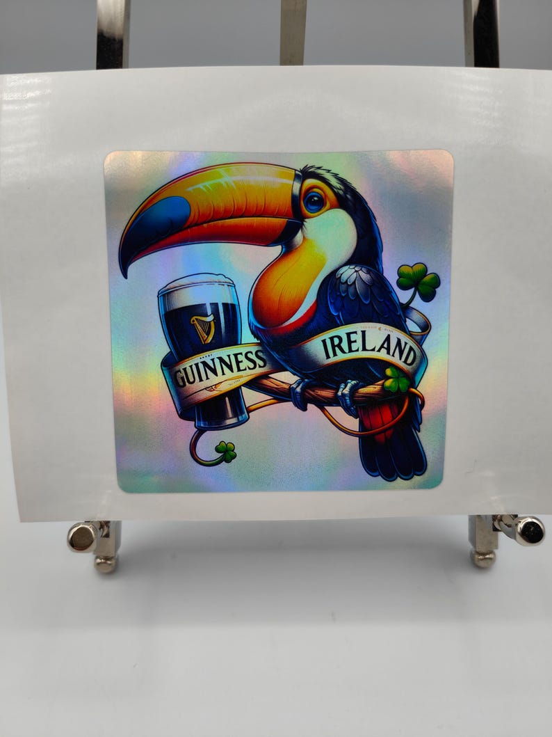 Op de afbeelding: Een holografische sticker met een toekan die een glas Guinness-bier vasthoudt. De toekan zit op een tak met een klavertjevier en het woord "IRLAND" op de tak. De sticker bevat ook de woorden "GUINNESS" en een klavertjevier.