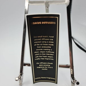 May include: A black and gold label with the text "ABACUS DIFFUSERS" and information about the hand-poured diffusers. The label is on a silver easel.