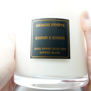 Könnte beinhalten: Ein klares Glas-Kerzengefäß mit einem schwarzen Etikett, auf dem "ABACUS PRINTS" und "60 mm x 60 mm" steht. Das Etikett listet auch die verfügbaren Farben auf: Gold, Silber, Blau, Rot, Kupfer, Schwarz.