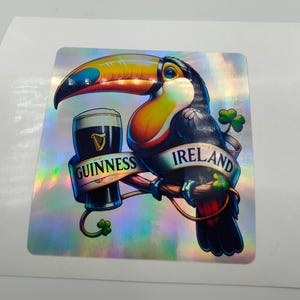 Op de afbeelding: Een toekan die een glas Guinness-bier vasthoudt met een banner waarop "Guinness Ireland" staat. De toekan zit op een tak met klavertjes eromheen. De achtergrond is een holografische, iriserende afwerking.