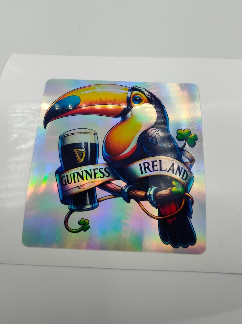 Op de afbeelding: Een toekan die een glas Guinness-bier vasthoudt met een banner waarop "Guinness Ireland" staat. De toekan zit op een tak met klavertjes eromheen. De achtergrond is een holografische, iriserende afwerking.