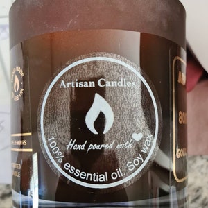 Op de afbeelding: Een bruine glazen kaars pot met een wit label waarop staat "Artisan Candles" en "Hand poured with 100% essential oil Soy wax".