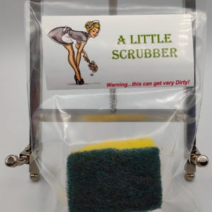 May include: A Little Scrubber sponge in a clear plastic bag, held by a metal stand. The sponge is yellow and green. The label features a pin-up style illustration and the text "A Little Scrubber" and "Warning...this can get very Dirty!"
