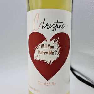 May include: A white wine bottle with a red heart and the text "Christine... Will You Marry Me?"  The heart has a scratch-off coating with the words "Scratch Me" below it.