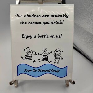 May include: A white sign with black text that reads "Our children are probably the reason you drink! Enjoy a bottle on us!"  The sign also features a blue banner with the text "From the O'Donnell family" and three stick figure children.
