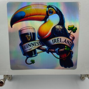 Op de afbeelding: Een toekan met een grote oranje snavel houdt een glas Guinness-bier vast. De toekan zit op een tak met klavertjes en het woord "IRLAND" op een banner. De banner onder de toekan zegt "GUINNESS".