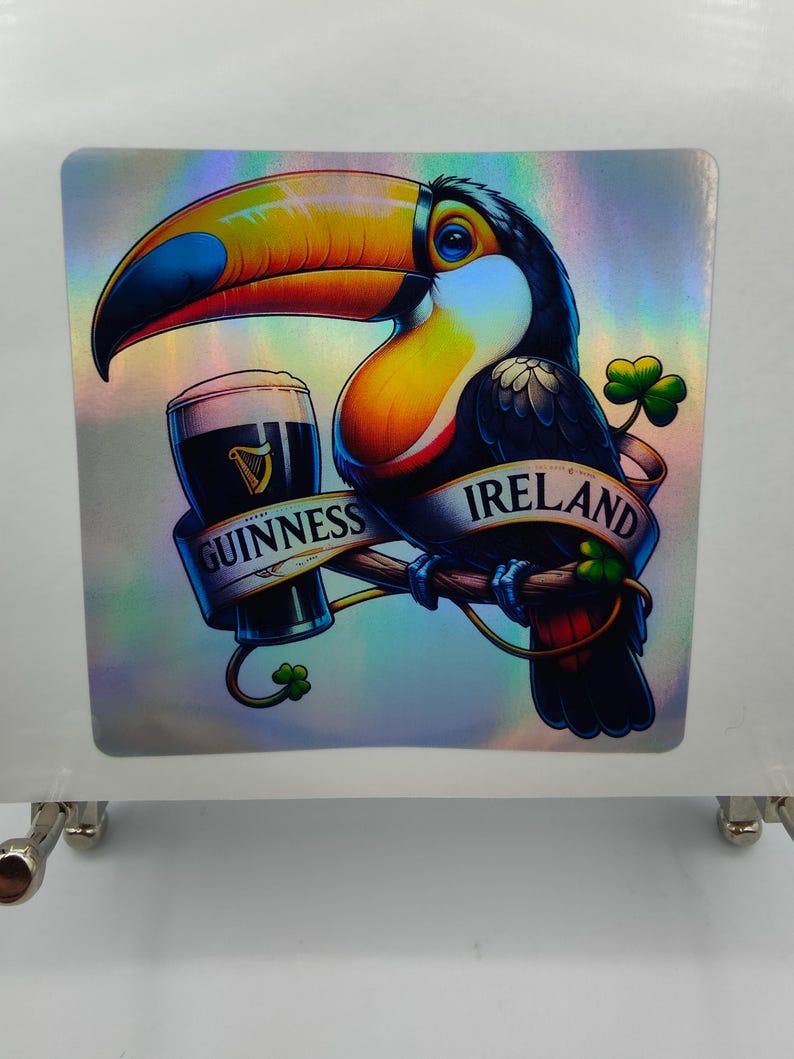 Op de afbeelding: Een toekan met een grote oranje snavel houdt een glas Guinness-bier vast. De toekan zit op een tak met klavertjes en het woord "IRLAND" op een banner. De banner onder de toekan zegt "GUINNESS".