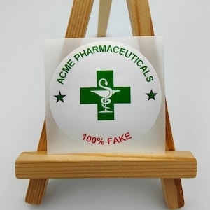 Może przedstawiać: Okrągła biała naklejka z zielonym krzyżem i wężem w środku. Tekst wok&oacute;ł krzyża brzmi "ACME PHARMACEUTICALS" z gwiazdą po obu stronach. Tekst pod krzyżem brzmi "100% FAKE" na czerwono.