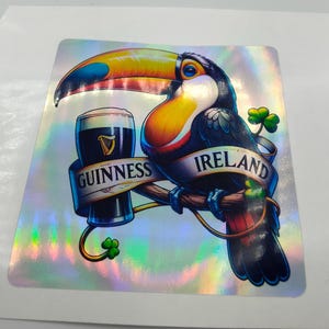 Op de afbeelding: Een toekan met een kleurrijke snavel en veren houdt een glas Guinness-bier vast. De toekan zit op een tak met een klavertjevier en de woorden "GUINNESS" en "IRLAND" op een banner.
