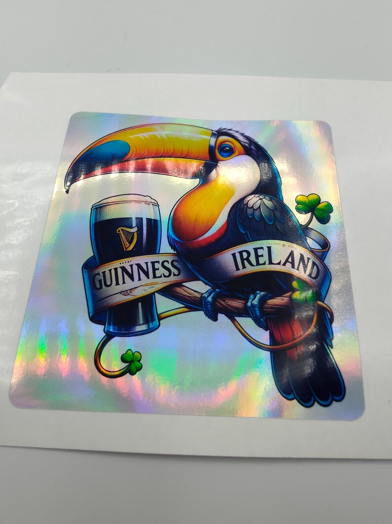 Op de afbeelding: Een toekan met een kleurrijke snavel en veren houdt een glas Guinness-bier vast. De toekan zit op een tak met een klavertjevier en de woorden "GUINNESS" en "IRLAND" op een banner.