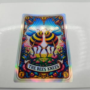 May include: A holographic card with two smiling bees, striped yellow and black, above a floral scene. The card reads "THE BEE'S KNEES" on a banner. The design is framed with decorative borders and details.