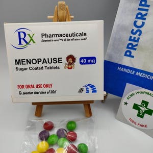 May include: A white box with the text "Rx Pharmaceuticals" and "Menopause Sugar Coated Tablets 40 mg" on it. The box is sitting on a small wooden easel. A bag with the text "Prescription" is in the background. A small bag of colourful sweets with the text "Skittles" is in the foreground.