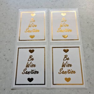 May include: Four white square stickers with gold borders and hearts. Each sticker has the text "Be Wise Sanitise" in gold lettering.
